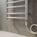Chrome towel radiator mounted on a tiled wall with a hose attached.